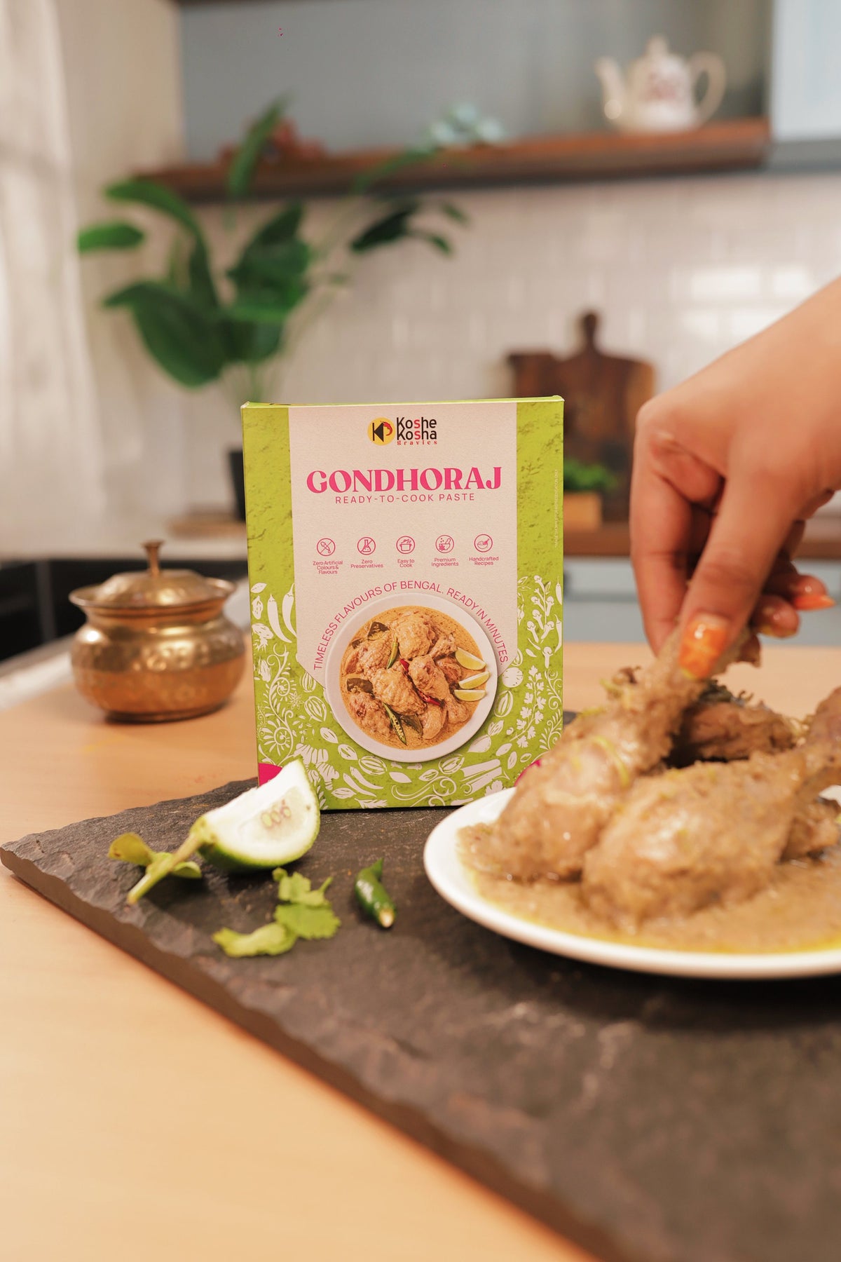 A hand lifting a piece of gondhoraj chicken from the plate, with the Gondhoraj box behind