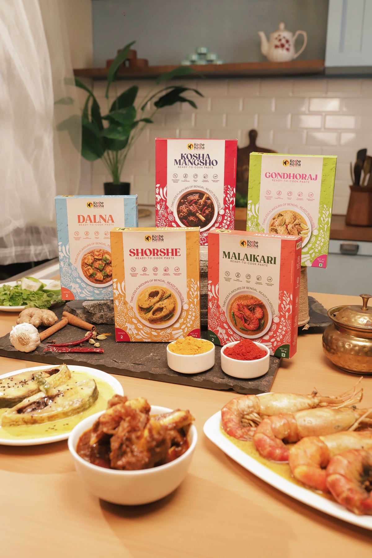 The five paste boxes on a kitchen counter behind plates of shorshe fish, mutton kosha and prawn malaikari
