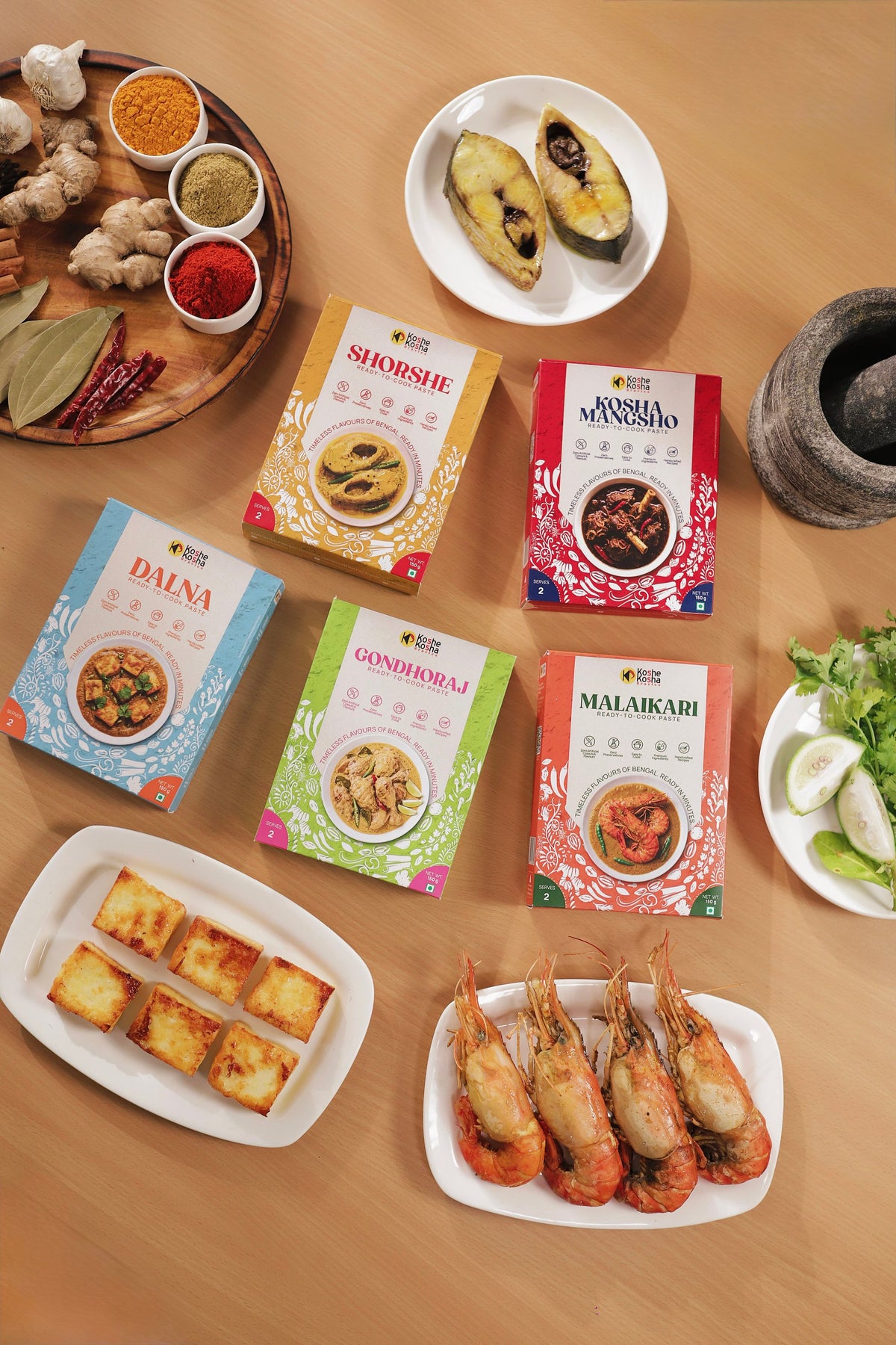 The five Koshe Kosha paste boxes on a wooden table with fish steaks, fried chhana, prawns and whole spices