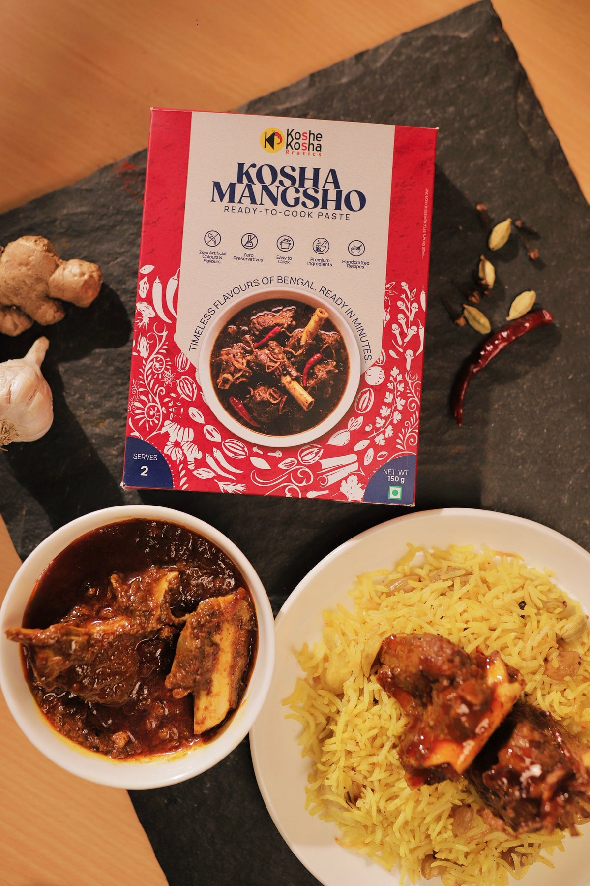 Kosha Mangsho paste box on black slate beside a bowl of dark mutton kosha and a plate of yellow pulao
