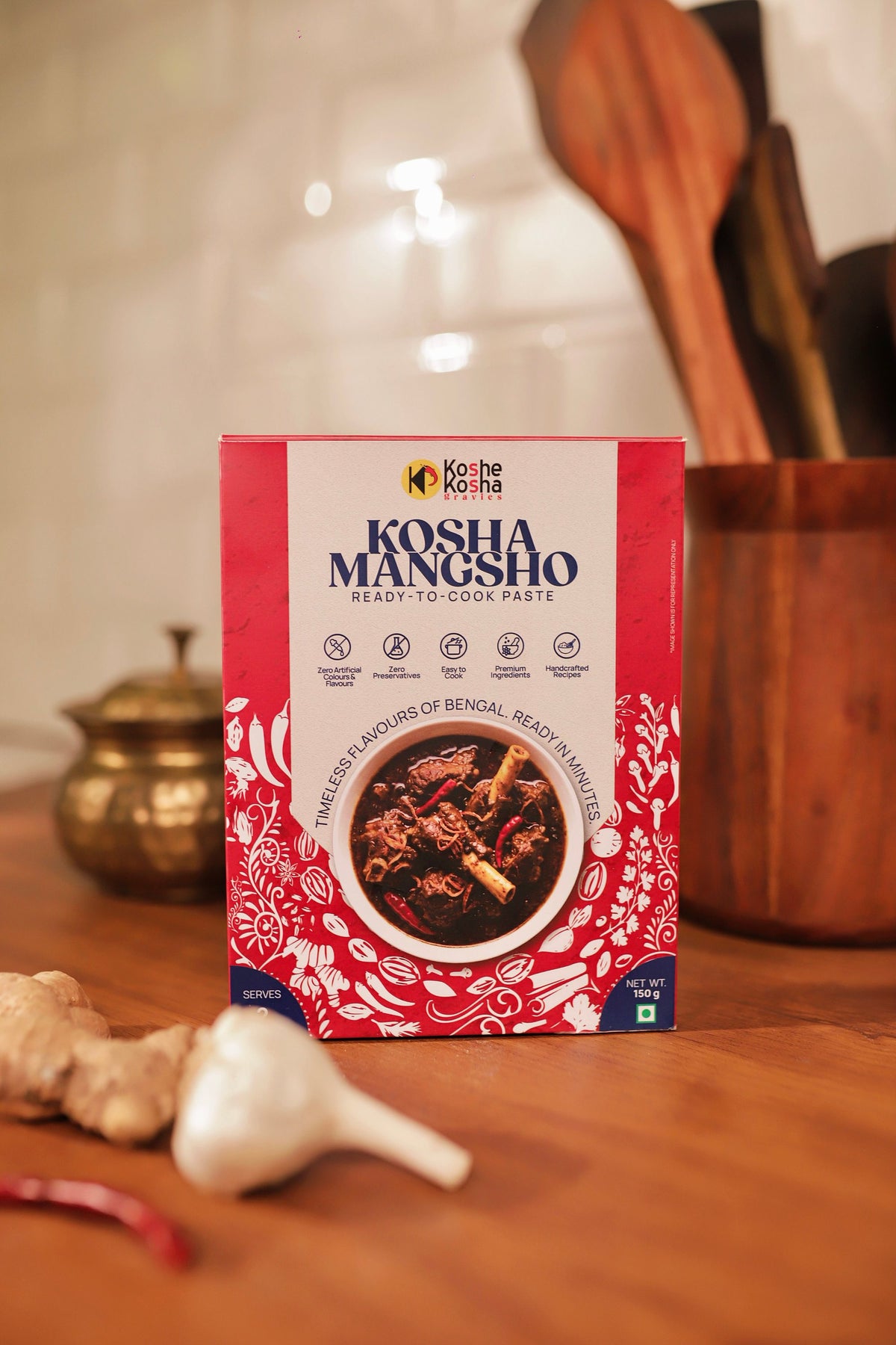Kosha Mangsho box on a wooden counter with a brass pot, garlic and wooden spoons