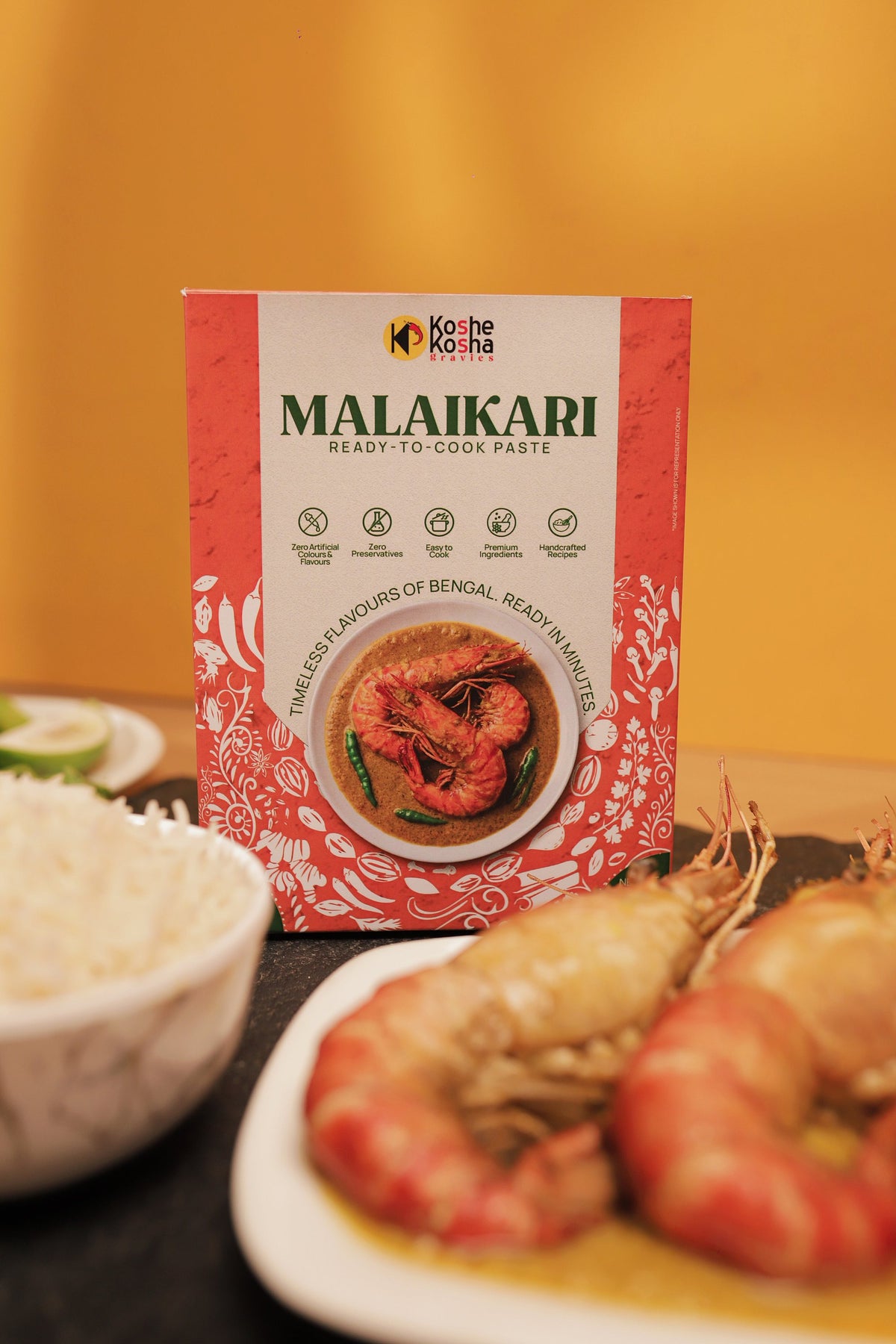 Malaikari box behind a plate of large prawns in malaikari gravy and a bowl of rice, against a yellow wall