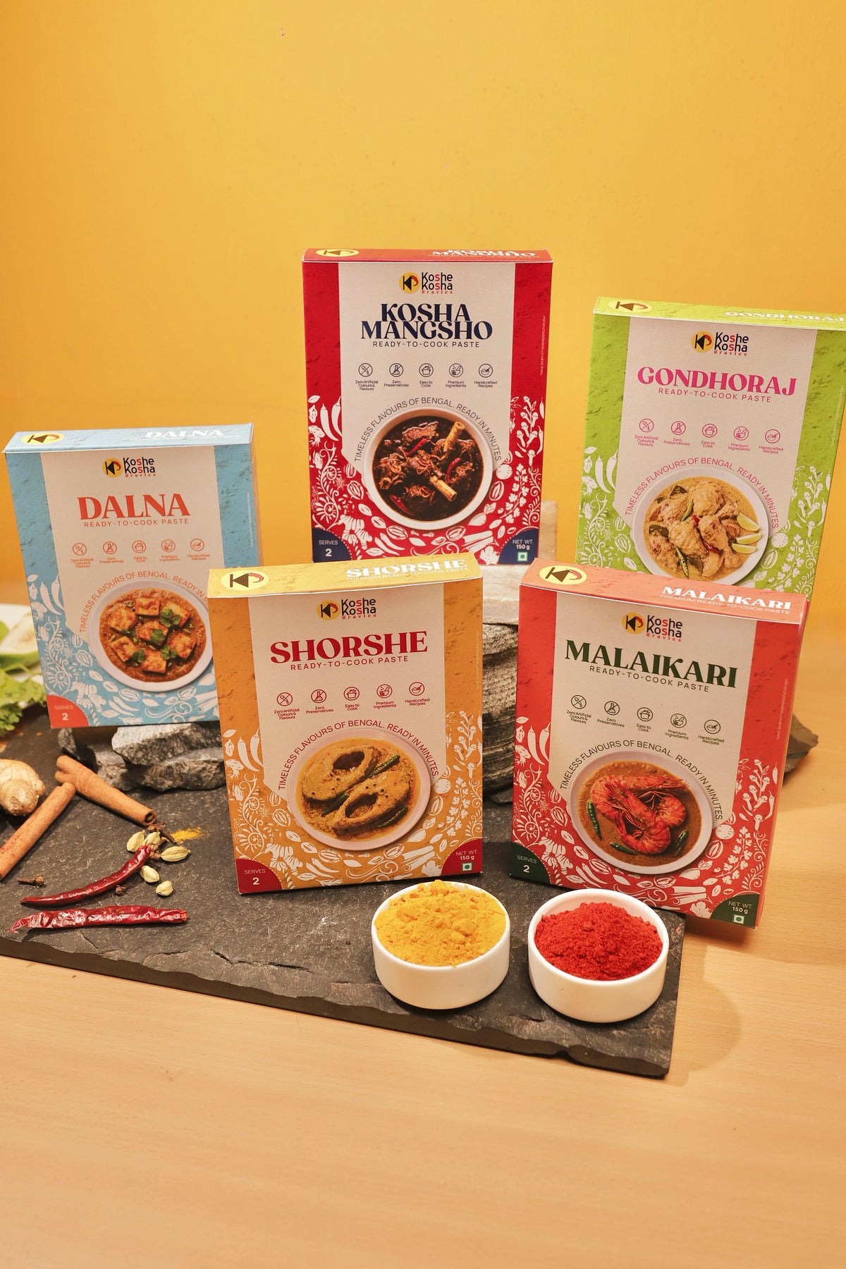 All five Koshe Kosha paste boxes on slate against a yellow wall, with bowls of turmeric and chilli powder