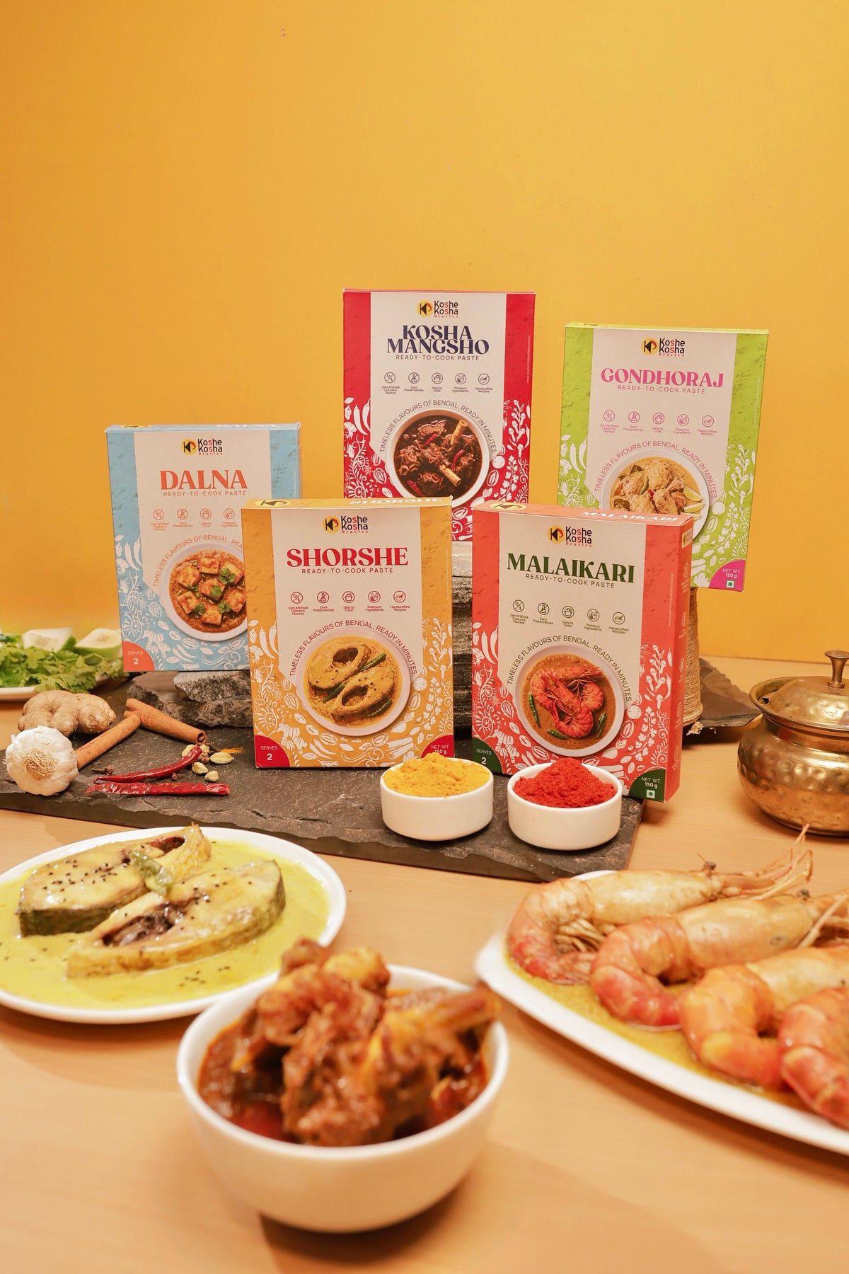 The five paste boxes behind plates of shorshe fish, mutton kosha and prawn malaikari, against a yellow wall